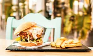 En hamburger med tilbehør av pommes frites på en tallerken, plassert på et bord med en stol i bakgrunnen. (AI-generert tekst)