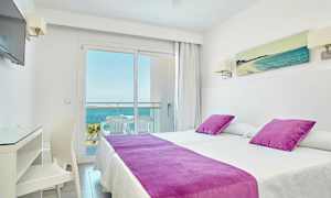 Et hotellrom med to senger kledd med hvite laken og lilla sengetepper og puter. En balkong med sitteplasser og utsikt mot havet er synlig gjennom en glassdør. (AI-generert tekst)