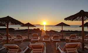 Solstoler på en strand med stråtakparaplyer i forgrunnen og en solnedgang over havet i bakgrunnen. (AI-generert tekst)