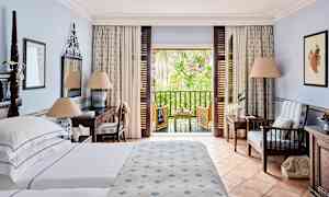 Hotellrom med seng, skrivebord og sitteområde samt en balkong med utsikt over grøntområder. (AI-generert tekst)