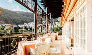 Restaurant med bord og stoler på en terrasse med utsikt over bygninger og fjell. (AI-generert tekst)