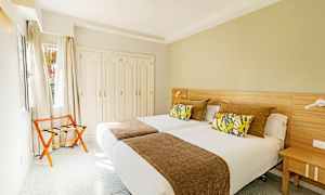 Et hotellrom med to enkeltsenger, dekorative puter og et brunt sengeteppe, samt en garderobe og en stol ved vinduet. (AI-generert tekst)