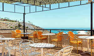 Utendørs servering med bord og stoler på en terrasse med utsikt over havet og en skråning med bygninger. (AI-generert tekst)