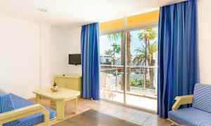 Et lyst hotellrom med en blåstripet sofa, et bord med en champagneflaske og glass, en liten TV på en møbel, og store vinduer med blå gardiner som leder ut til en balkong med utsikt mot palmer. (AI-generert tekst)