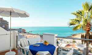 Utsikt fra en balkong med et bord dekket med en blå duk, omgitt av stoler. En kurv med frukt står på bordet, og i bakgrunnen sees stranden, palmer og havet. (AI-generert tekst)