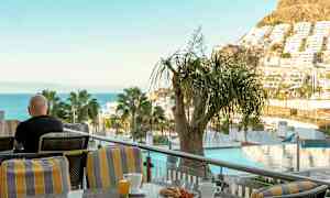 En person sitter på en terrasse med utsikt over havet, trær og bygninger, med en kopp kaffe og frokost foran seg. (AI-generert tekst)