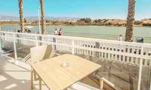 Utsikt fra en terrasse med bord og stol, med vann og gangvei i bakgrunnen. (AI-generert tekst)