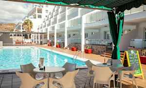 Utsikt over et svømmebasseng med solsenger og bord på terrassen, omgitt av bygninger. (AI-generert tekst)