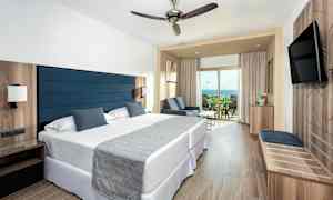 Et hotellrom med to senger, en sitteplass og en balkong med utsikt over havet. (AI-generert tekst)