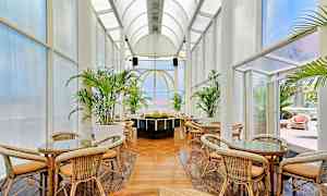 Kurvmøbler og bord under høyt hvelvet tak med store glasspartier omgitt av grønne planter. (AI-generert tekst)