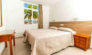 Et hotellrom med to enkeltsenger dekket med beige sengetepper. En vegg har et vindu som vender mot en palme og havet. (AI-generert tekst)