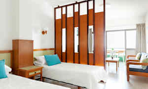 Et hotellrom med to enkeltsenger, et trebord og en sittegruppe avskilt av en romdeler. (AI-generert tekst)