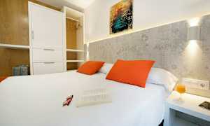 Soverom med hvit dobbeltseng, oransje puter og åpen garderobe. (AI-generert tekst)