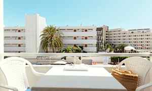 Utsikt fra en balkong med sittegruppe, bygninger og palmer synlige i bakgrunnen under en klar blå himmel. (AI-generert tekst)