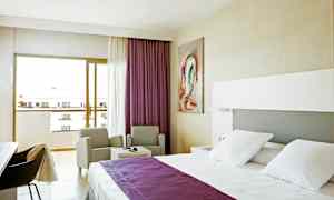 Et hotellrom med en seng med hvitt sengetøy og lilla overkast, to stoler ved et bord og en balkong med utsikt. (AI-generert tekst)