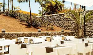 Utendørs restaurant med oppstilte bord og stoler, omgitt av steinmur og palmer. (AI-generert tekst)