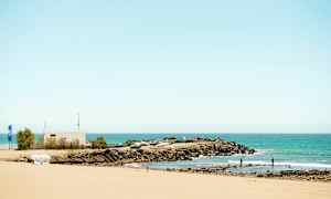 Sandstrand med utsikt over et klippeområde og småbåthavn ved havet. (AI-generert tekst)