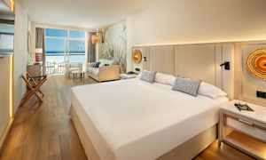 Et hotellrom med en stor seng, sofa og utsikt over havet gjennom vinduer. (AI-generert tekst)