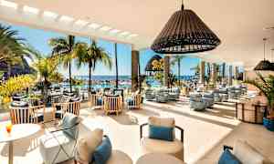 Interiør av en lys lounge med moderne møbler, store vinduer og utsikt over et bassengområde med palmer og sjø. (AI-generert tekst)