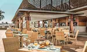 Et utendørs kaféområde med trebord, stoler og tak, dekorert med planter og duker, serverer mat og drikke. (AI-generert tekst)