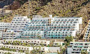 Bilde av et hotell med hvit fasade og balkonger, plassert ved en steinete skråning. (AI-generert tekst)