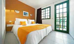 Et soverom med en dobbeltseng med hvite og oransje tekstiler, og store vinduer med grønne karmer. (AI-generert tekst)