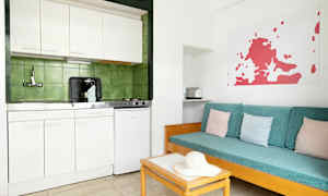 Et enkelt kjøkken med hvit innredning og grønne fliser bak. Ved siden av er det en sofa med turkise puter og pastellfargede puter. (AI-generert tekst)