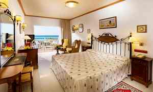 Et hotellrom med en dobbeltseng, sitteområde og en balkong med utsikt mot havet. (AI-generert tekst)