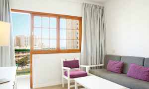 Et stue med grå sofa og stoler med lilla puter, og en tredør med glassdetaljer som viser en utsikt mot bygninger og havet. (AI-generert tekst)