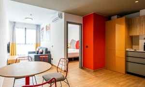 Et moderne innredet hotellrom med et bord og stoler i forgrunnen, en sittegruppe med sofa og et soverom synlig gjennom en døråpning. Kjøkkenområdet har skap og arbeidsflater. (AI-generert tekst)