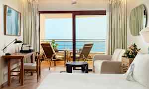 Et hotellrom med en balkong som har havutsikt. Rommet er møblert med skrivebord, stol, sofa, seng og blomster. (AI-generert tekst)