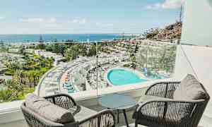 Utsikt fra en balkong over et hotell med et stort basseng, omkringliggende palmer og havet i bakgrunnen. (AI-generert tekst)