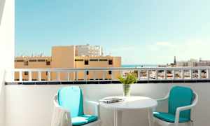 En balkong med to stoler, et bord og utsikt over bygninger og havet. (AI-generert tekst)