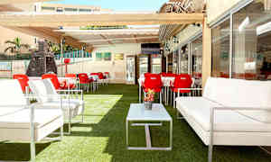 Utendørs servering med hvite sofaer, røde stoler og bord, plassert på kunstgress og med pergola. (AI-generert tekst)