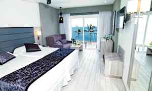 Hotellrom med dobbeltseng, sofa og havutsikt utenfor store vinduer. (AI-generert tekst)