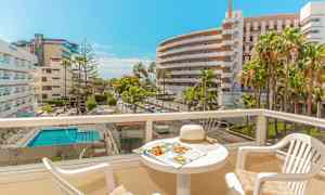 Utsikt over en solrik balkong med bord og stoler, hotellkompleks og basseng synlige i bakgrunnen. (AI-generert tekst)