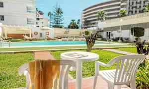 Et utendørs basseng omgitt av grøntområder og solsenger, med hvite bygninger i bakgrunnen. (AI-generert tekst)