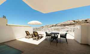 En terrasse med solsenger, parasoll og utemøbler, med utsikt mot sol og en fjellskråning. (AI-generert tekst)