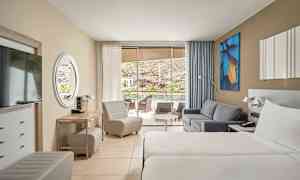 Hotellrom med to enkeltsenger, sofa og sittemøbler. Glasdører fører til en terrasse med utsikt. (AI-generert tekst)