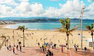 En strand med mennesker som spiller volleyball og går på strandpromenaden. Havet og fjellene vises i bakgrunnen. (AI-generert tekst)