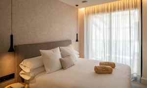 Et hotellrom med en stor seng, hvitt sengetøy og puter. På sengen ligger sammenrullede håndklær. Vegger og gardiner er lyse, og rommet har hengende lamper. (AI-generert tekst)