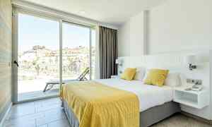 Et hotellrom med en stor seng dekket av hvitt og gult sengetøy. Rommet har et lyst og romslig preg med glassdører som leder ut til en balkong med utsikt over et fjellområde. (AI-generert tekst)