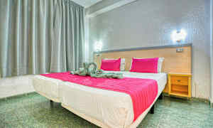 Seng med rosa teppe og puter i et rom med grå gardiner og en trebenk. (AI-generert tekst)