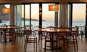 Restaurant med trestoler og bord med utsikt over havet og solnedgang. (AI-generert tekst)