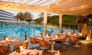 Utendørs restaurant med dekkede bord og lyslenker, plassert nær et basseng og omgitt av palmer og grøntområder. (AI-generert tekst)