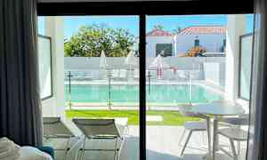 Utsikt fra et rom med stoler og bord mot et basseng med solsenger og parasoller utenfor. (AI-generert tekst)