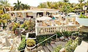 Utendørs restaurant med hvite parasoller og sitteplasser, omgitt av grøntområder og palmer. (AI-generert tekst)