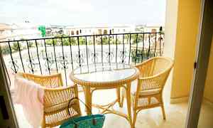 To rottingstoler og et rundt bord med glassplate står på en balkong med utsikt over steinbygninger og et tennisområde. (AI-generert tekst)