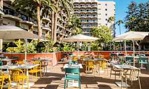 Bildet viser en utendørs restaurant med fargerike stoler, bord og parasoller foran et hotell med balkonger. (AI-generert tekst)