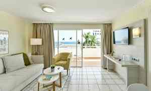 En stue med lyse møbler og en stor glassdør som fører til en balkong med utsikt over havet. (AI-generert tekst)